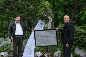 Nikolaos Marinakis und Aristomenis Syngelakis (mit Vorschlaghammer) installieren eine symbolische Gedenktafel für die Kriegsverbrechen Reichenhaller Gebirgsjäger vor dem Kreta-Gedenkstein in Bad Reichenhall. Im Hintergrund: Ein Kranz des AK Angreifbare Traditionspflege mit einer Meter langen Schleife, auf der die Orte von Massakern der deutschen Gebirgstruppe aufgeführt sind.